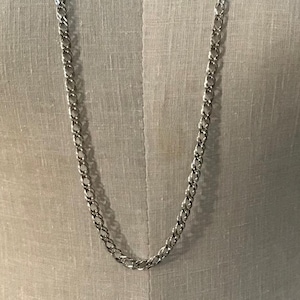 May include: A silver-colored chain necklace with a double-link design. The necklace is displayed against a neutral-toned background, showcasing its simple yet elegant style. The chain is made of a shiny metal.