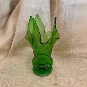 May include: A green glass vase with a unique, flared design. The vase has a textured surface and a wide, open top with pointed edges. The base is round and has a pedestal. The vase is approximately 8 inches tall.