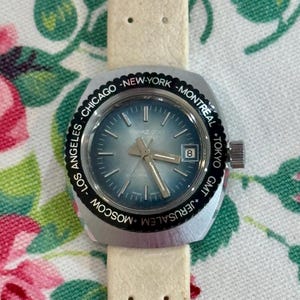 Puede incluir: Un reloj vintage plateado con esfera azul claro y correa de cuero crema. La esfera del reloj muestra el texto "TOPAZ 200" y la fecha. El bisel negro está inscrito con nombres de ciudades como Los Ángeles, Chicago y Nueva York.
