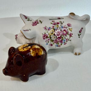 Puede incluir: Dos cerditos de cerámica. Uno es blanco con diseños florales en rosa, amarillo y azul. El otro es un cerdito pequeño, marrón y vidriado. Ambos tienen ranuras para insertar monedas.