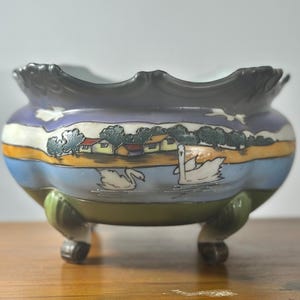 Puede incluir: Un cuenco de cerámica decorativo con una escena de paisaje que representa cisnes, casas y árboles. El cuenco tiene un borde festoneado y descansa sobre tres pequeños pies. La paleta de colores incluye azul, verde, amarillo y blanco.