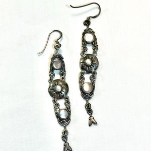 May include: A pair of dangling earrings featuring silver-toned metal with an oxidized finish. Each earring consists of three circular settings containing iridescent white cabochons, with a sunburst design on the center piece and a small charm.