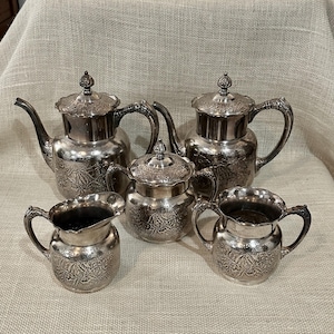 May include: A silver tea set with ornate floral detailing. The set includes two teapots, a sugar bowl with a lid, and two creamers. The pieces have curved handles and decorative finials on the lids. The set is displayed on a neutral-toned surface.