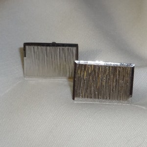 May include: Two rectangular silver cufflinks with a textured, vertical line pattern. The cufflinks feature a polished metal frame and a reflective surface, giving a modern and sophisticated appearance. The background is a light, neutral fabric.