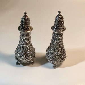 May include: A pair of silver salt and pepper shakers with an ornate floral design.