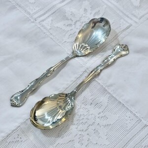 Puede incluir: Dos cucharas de servir plateadas ornamentadas con cuencos en forma de concha y mangos detallados. Las cucharas se exhiben sobre un mantel de encaje blanco, reflejando la luz. Las cucharas tienen una estética vintage.