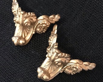 Könnte beinhalten: Zwei goldfarbene Eckkonsolen aus Metall mit verzierten floralen Designs. Die Konsolen sind rechtwinklig geformt und haben ein dekoratives, rollenförmiges Design.