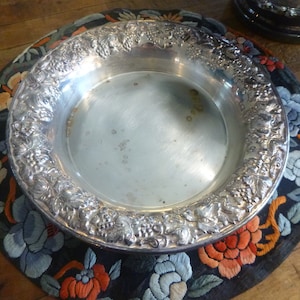 May include: A round silver-toned metal bowl featuring a wide, ornate rim decorated with embossed grape clusters and vine leaf patterns. The interior of the bowl has a smooth, reflective surface with visible small brown spots and light tarnishing.