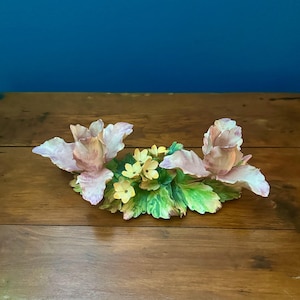 May include: A decorative porcelain floral arrangement featuring two large, pale pink and white roses positioned on opposite ends, connected by a central cluster of small yellow blossoms and detailed green leaves with visible veining.