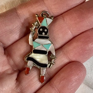May include: A colorful pendant in the shape of a Native American dancer. The pendant features a silver-tone metal frame and is inlaid with various colors, including red, turquoise, black, and white. The dancer is holding a staff.