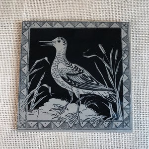 May include: Black and white tile featuring a stylised image of a bird standing on a bed of reeds. The bird has a long beak and a detailed pattern of feathers. The tile has a decorative border.