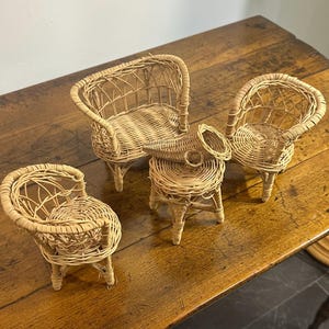 May include: Four miniature wicker furniture pieces: a small sofa, two armchairs, and a stool. The furniture is made of light brown woven wicker and sits on a wooden surface. The pieces are designed for a dollhouse or decorative display.