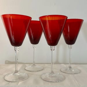 Op de afbeelding: Een set van vier bijpassende wijnglazen met dieprode, conisch gevormde kelken en heldere glazen stelen met ronde voeten. De glazen hebben een gladde, brede rand en een taps toelopend ontwerp dat smaller wordt naar de basis van de kelk.