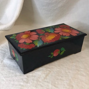 May include: A black wooden box with a hinged lid. The lid is painted with a floral design featuring red, pink, yellow, and green flowers. The box has a rectangular shape and a slightly raised base.