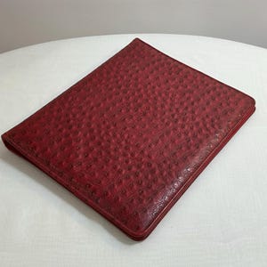 May include: A red leather portfolio with a textured, dotted pattern. The rectangular case is closed and lies flat on a white surface. The edges of the portfolio are slightly darker than the main body.