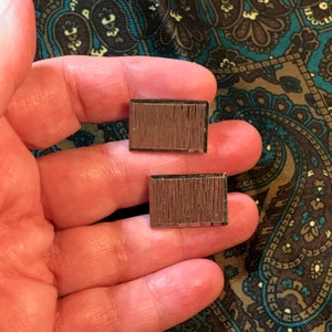 May include: Two rectangular cufflinks with a textured, brown surface. The cufflinks have a dark border and are displayed against a patterned background. These accessories are suitable for formal wear.