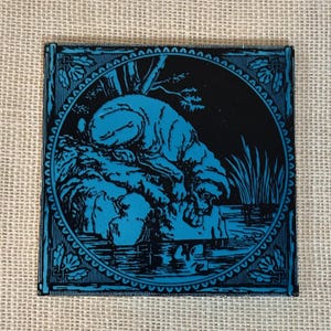 May include: A blue and black tile with a decorative border. The tile features a black and white illustration of a dog lying down by a body of water.