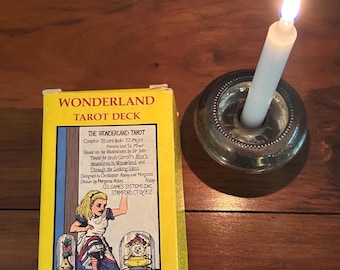 Pode incluir: Uma caixa amarela "Wonderland Tarot Deck" com ilustrações de Alice no País das Maravilhas está ao lado de uma vela branca acesa em um castiçal de prata. A caixa tem texto e ilustrações.