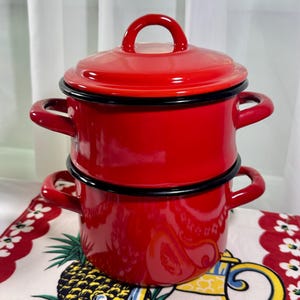 Puede incluir: Una olla doble de esmalte rojo brillante con dos asas laterales en cada sección y una tapa roja a juego. La olla presenta un acabado brillante con un borde negro distintivo alrededor de la parte superior de la base y la tapa.