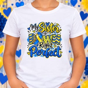 May include: A white short-sleeved t-shirt featuring a graphic design with the text My Sister is Down Right Perfect. The lettering is in blue and yellow with a ribbon symbol integrated into the word Down, surrounded by small blue and yellow hearts.