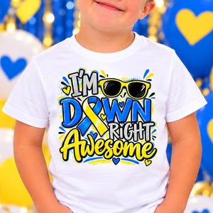 May include: A white short-sleeved t-shirt featuring a graphic print with the text I'M DOWN RIGHT AWESOME in blue, yellow, and black lettering, accompanied by an illustration of black sunglasses and a blue and yellow awareness ribbon.