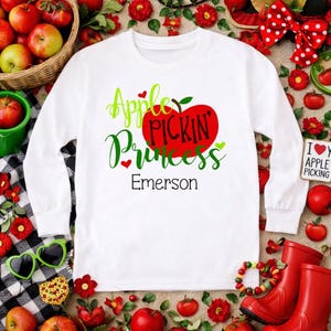 May include: White long-sleeved shirt with the text "Apple Pickin' Princess" and the name "Emerson" in green and black. The design includes a red apple graphic. The shirt is surrounded by apples, flowers, and red boots.