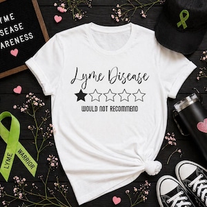 May include: White t-shirt with the words "Lyme Disease" and a rating of one star out of five, with the text "Would Not Recommend". A black cap with a green ribbon, a green ribbon with the words "Lyme Warrior", and black sneakers are also visible.