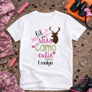 May include: White t-shirt with the text "lil Miss Camo cutie" in pink and green, a brown deer silhouette, and the name "Emalyn" in black. The shirt is surrounded by pink camouflage clothing, pink accessories, and black boots.