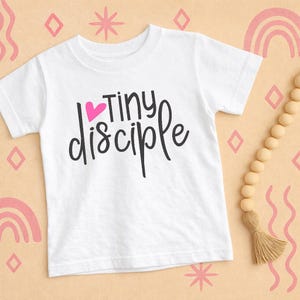 May include: White t-shirt with the text "Tiny Disciple" in black script, with a pink heart above the word "Tiny". The shirt is displayed on a light peach background with decorative shapes and a wooden bead garland.