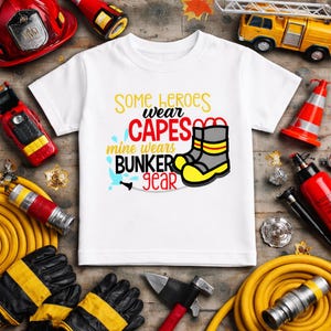 May include: White t-shirt with the text "Some heroes wear capes, mine wears bunker gear" in yellow, red, and blue. The shirt features a graphic of firefighter boots. Firefighter-themed items surround the shirt, including a helmet, gloves, and a fire truck.