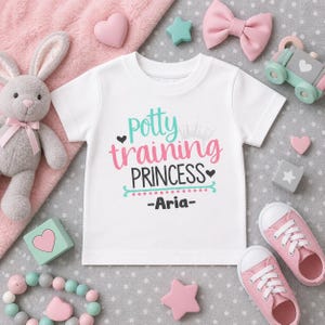 May include: White t-shirt with the text "Potty Training Princess - Aria-" in teal and pink. The shirt is surrounded by baby toys, including a stuffed bunny, pink bow, and pink sneakers.