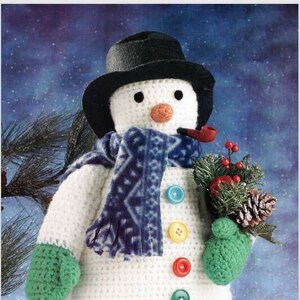 May include: A crocheted snowman wearing a black hat, blue scarf, and green mittens. The snowman has a red pipe in its mouth and is holding a small pine branch with red berries. The snowman is standing on a white surface with red, green, and blue ornaments.