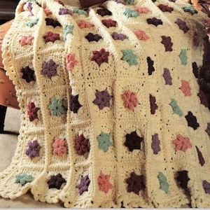 以下が含まれることがあります： クリーム色の背景にカラフルな花柄が施された、かぎ針編みの毛布。毛布は、紫、ピンク、緑、茶色のさまざまな色合いで作られています。