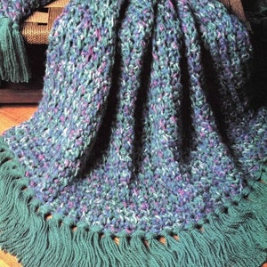 May include: A crocheted throw blanket with a multi-coloured yarn in shades of blue, green, and purple. The blanket has a fringe border in a solid green colour.