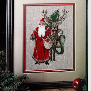 May include: Framed cross-stitch artwork featuring Santa Claus in a red coat and hat, with a white beard, walking beside a reindeer. The reindeer has a green Christmas tree on its back. The artwork is framed in a brown wooden frame.