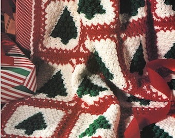 May include: A crocheted blanket with a red, white, and green Christmas tree pattern. The blanket is folded and draped over a stack of wrapped gifts.