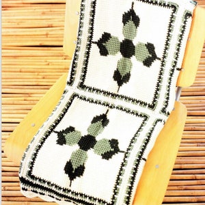 May include: A white crocheted throw blanket with a black and green geometric pattern. The blanket is folded and placed on a wooden chair.