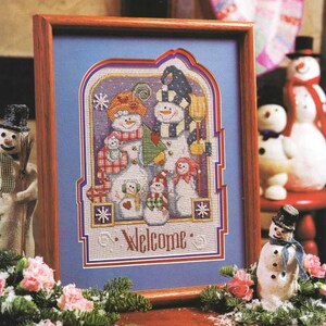 May include: A framed cross-stitch featuring a family of snowmen. The snowmen wear hats, scarves, and mittens, and hold a broom and a gift. The word "Welcome" is stitched at the bottom. The frame is wood.