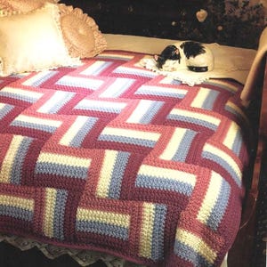 May include: A handmade crochet blanket with a geometric design in burgundy, pale blue, and cream. The blanket is on a bed, with pillows and a decorative cat figurine.