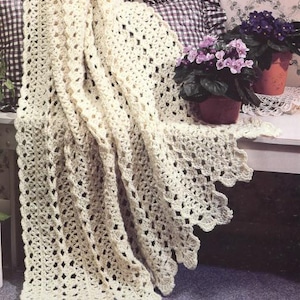 May include: A crocheted blanket in a cream color with a scalloped edge. The blanket is draped over a bench with a checkered pillow and a potted plant with purple flowers.