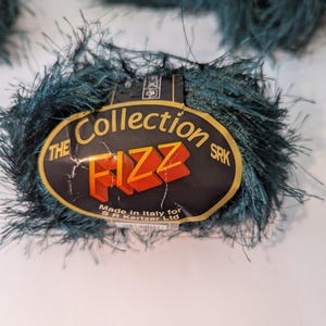 May include: Close-up of teal-colored yarn balls. The yarn has a fuzzy texture and a label that reads "The Collection Fizz SRK Made in Italy for S.R. Kortzer Ltd.". The label is black with gold and orange text. The yarn is likely for knitting or crochet projects.