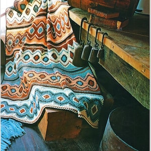 May include: A crocheted blanket with a geometric pattern in shades of blue, orange, and brown. The blanket is draped over a wooden stool and a wooden table with a barrel and bells.