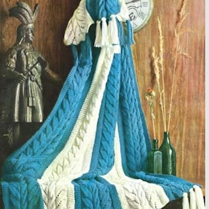 以下が含まれることがあります： ケーブル編みのデザインとタッセルが付いた、青と白のニットブランケット。木製の表面に掛けられており、背景には時計や装飾品があります。ブランケットの寸法は表示されていません。