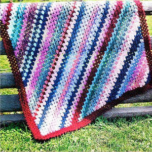 May include: A colorful crocheted blanket with a diagonal pattern in shades of blue, pink, white, and red. The blanket is draped over a wooden fence.