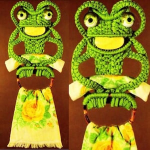 May include: Green crocheted frog towel ring with a yellow smile and large eyes. The ring is holding a yellow floral patterned towel.