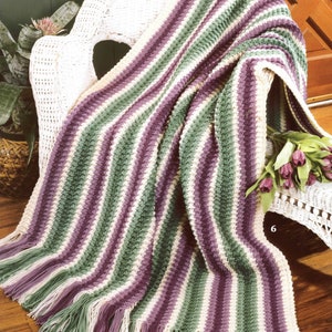 May include: A striped crocheted throw blanket in shades of purple, green, and white with a fringe border. The blanket is draped over a white wicker chair.