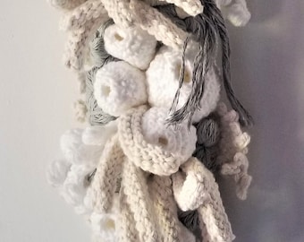 May include: A wall-hanging fibre art piece composed of various crocheted elements in shades of white, cream, and grey. The structure features numerous spherical, hollow-centred forms, long dangling yarn strands, and textured, organic-shaped clusters.