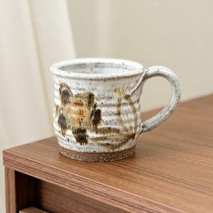 Puede incluir: Taza de cerámica hecha a mano con esmalte blanco y diseño floral marrón. La taza tiene un cuerpo redondeado, un asa y una superficie texturizada. Mide aproximadamente 10 cm de alto y 8 cm de diámetro, perfecta para café o té.