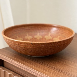 Può includere: Una ciotola rotonda in ceramica marrone con una superficie strutturata e un motivo floreale decorativo. La ciotola è esposta su una superficie di legno, mettendo in risalto il suo design rustico. Questa ciotola artigianale è adatta per servire cibo o come elemento decorativo.