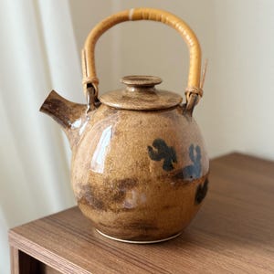 May include: A brown ceramic teapot with a bamboo handle. The teapot has a rounded shape, darker brown accents, and a small, dark blue floral design. The lid has a small knob, and the spout is angled. A decorative kitchen item.
